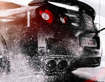 Detalle del prelavado con pistola a presión en Lavado Suave Hanna, aplicando agua a presión sobre la parte trasera y tubos de escape de un coche deportivo negro.